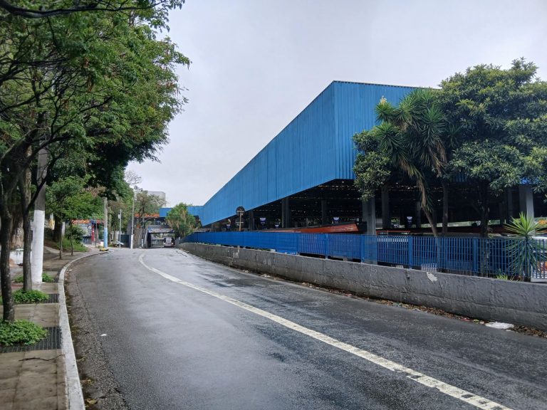 Aparecida de Goiânia credencia empresas para distrito com projeção de 20 mil empregos Estrada ao lado de um grande galpão industrial azul em um dia nublado.